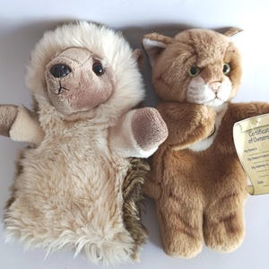 May include: Two plush hand puppets: a hedgehog with a cream-colored fur mane and a tan cat with green eyes. The cat puppet has a tag that reads "Certificate of Ownership". Both puppets are on a white background.