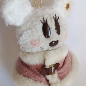 May include: A plush white mouse-shaped keychain with a pink and white coat. The mouse has brown eyes and eyelashes, pink cheeks, and a brown mouth. A gold chain is attached to the top of the mouse.