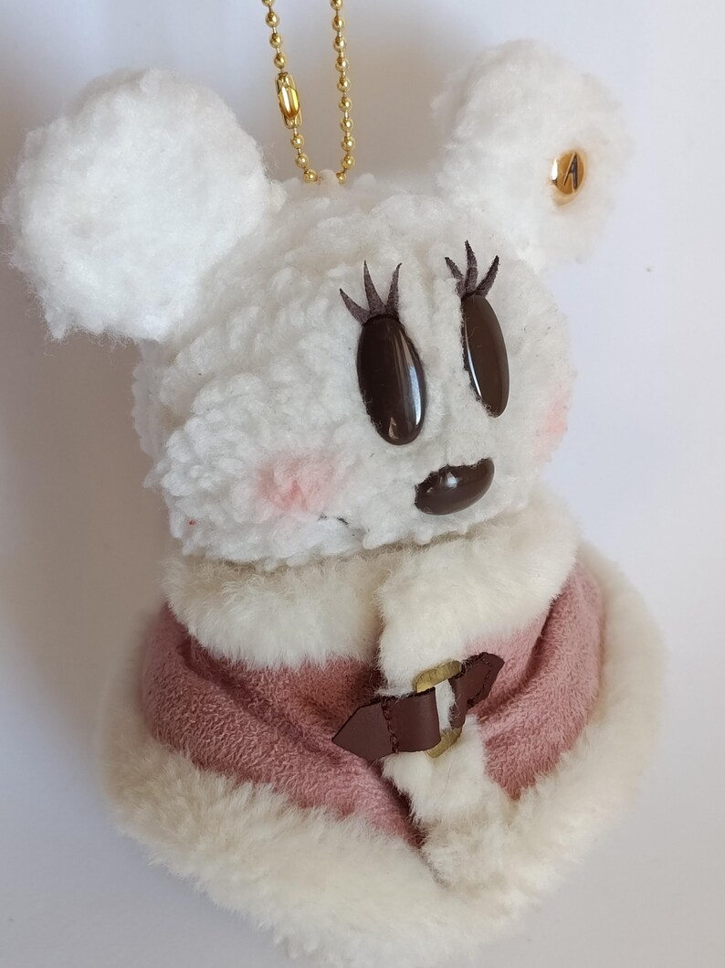 May include: A plush white mouse-shaped keychain with a pink and white coat. The mouse has brown eyes and eyelashes, pink cheeks, and a brown mouth. A gold chain is attached to the top of the mouse.