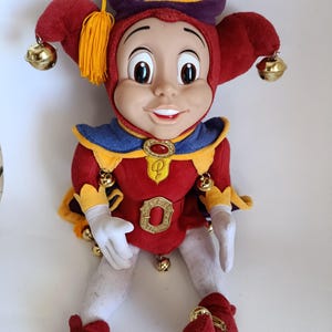 May include: A plush jester doll with a vinyl face, wearing a red and purple hat with a gold star and yellow tassel. The doll features a red tunic with a blue collar, a gold belt buckle, white gloves, white leggings, and red shoes, all accented with small gold bells.