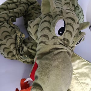 Pode incluir: Um dragão de pelúcia com corpo verde-claro e padrão de escamas em forma de meia-lua verde-escura, olhos grandes brancos e pretos, língua vermelha brilhante e uma asa de tecido dourado metálico. O corpo longo e flexível está enrolado.