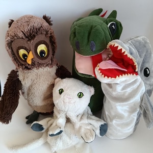 May include: A collection of four plush hand puppets including a brown owl with large yellow eyes, a green dragon with a pink tongue, a white cat with green eyes, and a gray shark with an open mouth showing white teeth and a red interior.