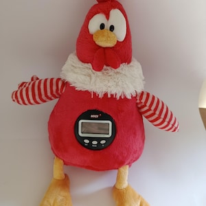 May include: A plush red rooster-shaped alarm clock with a digital display. The rooster has white-rimmed eyes, a yellow beak, and a white fluffy collar. Its arms are red and white striped, and it has yellow feet. The clock face reads "NICI".