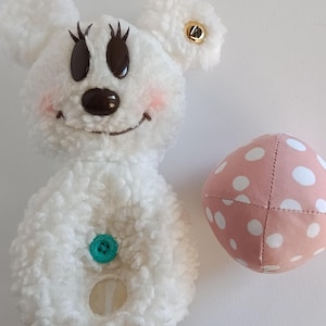 May include: A white plush toy shaped like a cartoon mouse with black eyes, eyelashes, and a brown nose. The mouse has pink cheeks, a green button, and a gold button on one ear. A pink polka-dot ball is next to the mouse.