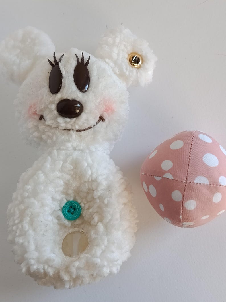 May include: A white plush toy shaped like a cartoon mouse with black eyes, eyelashes, and a brown nose. The mouse has pink cheeks, a green button, and a gold button on one ear. A pink polka-dot ball is next to the mouse.