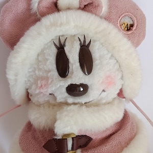 May include: A plush toy featuring a character with a white fur face, brown oval eyes, and a brown nose. The character wears a pink hat with matching ears and a pink and white fur-trimmed coat. A gold-colored clasp secures the coat.