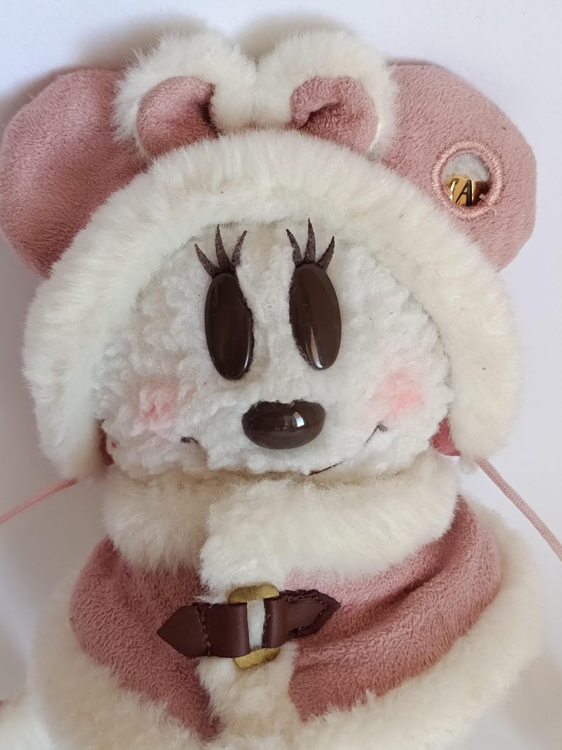 May include: A plush toy featuring a character with a white fur face, brown oval eyes, and a brown nose. The character wears a pink hat with matching ears and a pink and white fur-trimmed coat. A gold-colored clasp secures the coat.