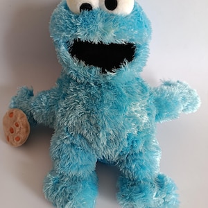 May include: A plush Cookie Monster toy, featuring a light blue, shaggy texture. The toy has large white eyes with black pupils, a wide open mouth, and a chocolate chip cookie in one hand. The toy is a popular character from a children's television show.