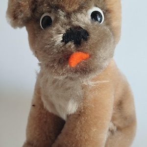 Puede incluir: Un perro de peluche marrón y blanco con grandes ojos negros y una lengua naranja que sobresale. El perro está sentado con las patas metidas debajo del cuerpo.