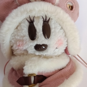 May include: A plush toy featuring a cartoon character with a white, fluffy face, brown eyes, and a small brown nose. The character wears a pink coat with a brown buckle and a matching pink hat with a bow. The cheeks are lightly blushed.