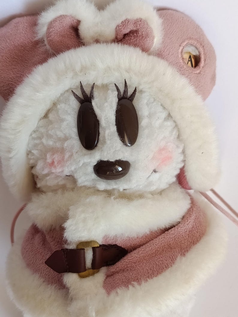 May include: A plush toy featuring a cartoon character with a white, fluffy face, brown eyes, and a small brown nose. The character wears a pink coat with a brown buckle and a matching pink hat with a bow. The cheeks are lightly blushed.