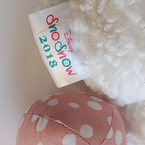May include: A close-up of a plush toy with a white, textured body and a pink polka-dotted foot. A tag reads "Snow 2018 Disney Snow" in red and teal lettering.