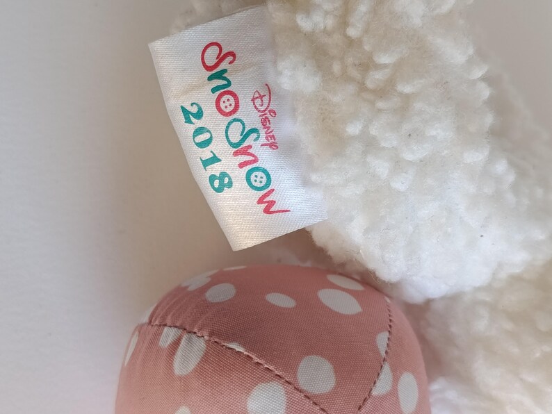 May include: A close-up of a plush toy with a white, textured body and a pink polka-dotted foot. A tag reads "Snow 2018 Disney Snow" in red and teal lettering.