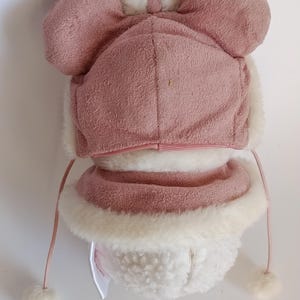 May include: A pink and white winter hat with ear flaps and a matching scarf. The hat features a bow on top and pom-poms on the strings. The hat and scarf are made of a soft, plush material, perfect for keeping warm in cold weather.