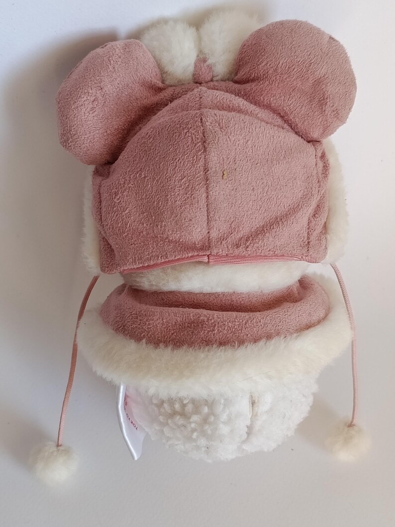 May include: A pink and white winter hat with ear flaps and a matching scarf. The hat features a bow on top and pom-poms on the strings. The hat and scarf are made of a soft, plush material, perfect for keeping warm in cold weather.