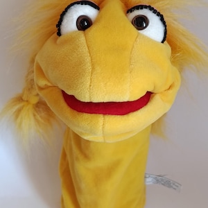 Op de afbeelding: Een gele pluche handpop met een zachte stoffen textuur, felgeel haar in zijvlechten, grote witte ogen met zwarte pupillen en dikke zwarte wimpers, en een brede mond met een rode binnenvoering.