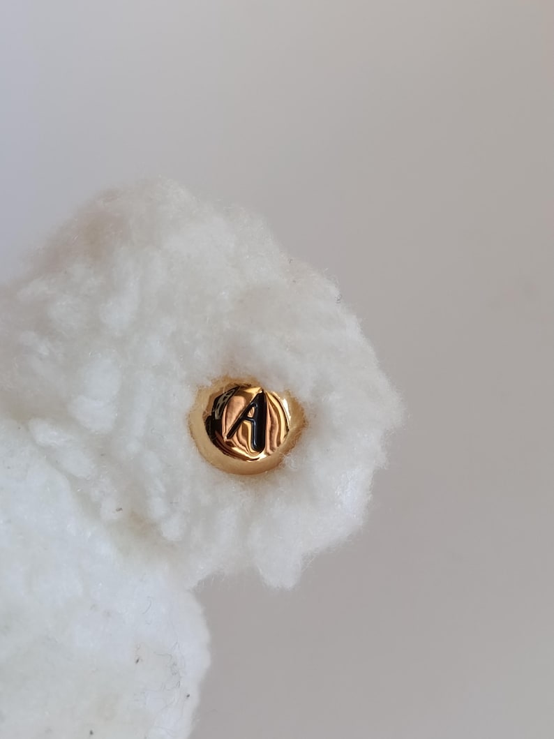 May include: Close-up of a gold-colored button with the letter "A" embossed on it, set against a backdrop of fluffy white material. The button has a shiny, reflective surface. The white material has a soft, cloud-like texture.