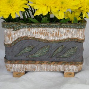May include: A rectangular ceramic planter with a distressed white, gray, and brown finish, filled with vibrant yellow chrysanthemums. The planter features a decorative band with leaf imprints and sits on small feet.