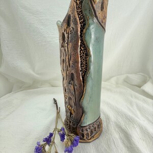 Puede incluir: Un jarrón de cerámica hecho a mano con un diseño único. El jarrón presenta una combinación de texturas y colores, incluyendo marrón, verde y dorado. El jarrón tiene un patrón calado texturizado en un lado y una superficie lisa de color verde claro en el otro. Una ramita de flores moradas descansa cerca del jarrón.