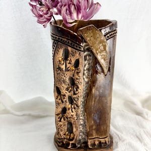 Puede incluir: Un jarrón de cerámica hecho a mano con un diseño único. El jarrón presenta una superficie texturizada de color bronce con detalles en marrón oscuro y un patrón decorativo. Dos flores moradas secas se colocan dentro del jarrón. El jarrón mide unos 20 cm de alto.