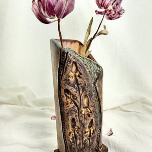 Puede incluir: Un jarrón de cerámica alto y rectangular con un exterior texturizado de color bronce y una base marrón oscuro. El jarrón presenta diseños florales en relieve y contiene dos tulipanes morados secos. El jarrón mide aproximadamente 25 cm de alto.