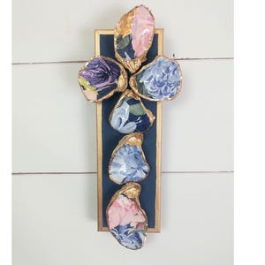 May include: A decorative wall hanging featuring five oyster shells with floral patterns in pink, blue, and purple. The shells are mounted on a navy blue rectangular frame with gold trim, creating a coastal or nautical theme.