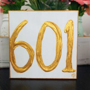 May include: A white canvas sign with the number 601 painted in gold. The number is painted in a thick, textured style.
