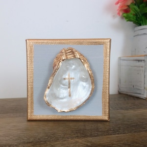 May include: A white oyster shell with a gold cross painted inside, mounted on a gray canvas with a gold frame.