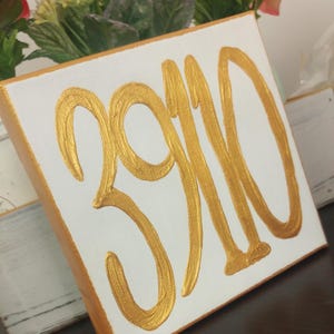 May include: A white canvas with the number 3910 painted in gold. The number is written in a large, stylized font.