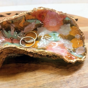 May include: A decorative oyster shell ring dish with a gold-painted exterior and colorful interior. The interior features a mix of orange, pink, and green hues. Two silver rings and a small turquoise object rest inside the shell.