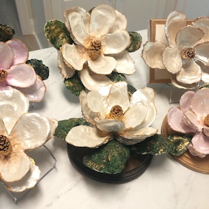 May include: A collection of handcrafted magnolia flower sculptures. The flowers feature creamy white and pink petals with gold accents, each with a pinecone center. The green leaves have gold detailing. Some are mounted on wooden bases.