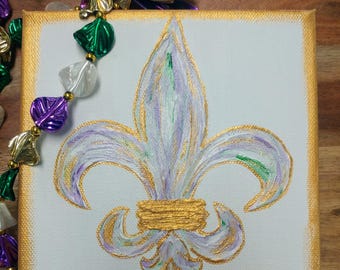May include: A white canvas painting of a purple and gold fleur-de-lis on a white background. The fleur-de-lis is painted with a textured brushstroke effect.