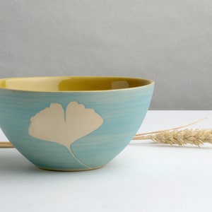 Pode incluir: Uma tigela de cerâmica com exterior azul claro e interior amarelo. Um desenho de folha de ginkgo creme na lateral. A tigela está sobre uma superfície branca com espigas de trigo.