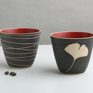 May include: Two black ceramic mugs with red interiors. One mug features a white ginkgo leaf design, the other has a white wavy line design.
