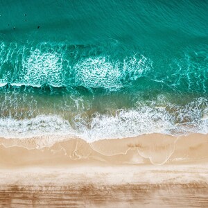 May include: Aerial view of a beach with turquoise water and sandy shore. White foamy waves crash onto the beach. Several dark figures are in the water. The sand is light brown and the water is a deep turquoise color.