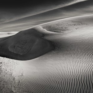 May include: A black and white photograph of sand dunes with rippled textures. The dunes are in the foreground and middle ground, with a cloudy sky in the background. The image is a landscape photograph.