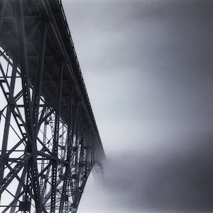 May include: A black and white photo of a metal bridge structure disappearing into a thick fog. The bridge is made of steel beams and girders, creating a geometric pattern.