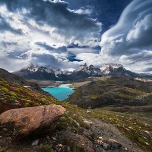 Op de afbeelding: Een schilderachtig landschap met besneeuwde bergen onder een dramatische, bewolkte lucht. Een turquoise meer ligt aan de voet van de bergen, omringd door groene heuvels en een grote, bruine rots op de voorgrond. De lucht is gevuld met wervelende wolkenformaties.
