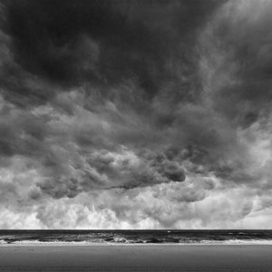 May include: A black and white photograph of a stormy sky over a beach. The sky is filled with dark, dramatic clouds, and the beach is empty.