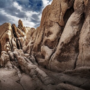 May include: A landscape photograph of a desert canyon with large, textured rock formations in shades of brown and tan. The sky is overcast with dark clouds, creating a dramatic contrast. A person is visible in the distance.