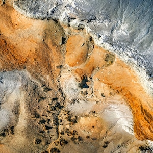 May include: Aerial view of a landscape with a mix of textures and colors. The image features areas of gray, orange, and brown, creating a visually interesting pattern. The textures vary from smooth to rough, suggesting a geological formation.
