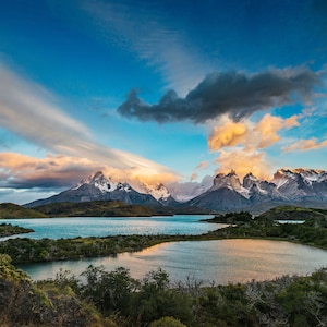 May include: A panoramic view of a mountain range with snow-capped peaks reflected in a still lake. The sky is a vibrant blue with fluffy white clouds and a long, thin cloud stretching across the sky.