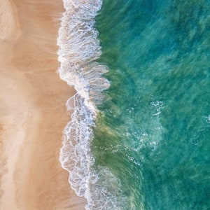 May include: Aerial view of a sandy beach meeting turquoise ocean water with two people paddleboarding.