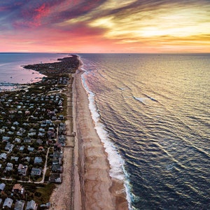 May include: Aerial view of a long, narrow island with a town on one side and a sandy beach on the other. The sky is a vibrant mix of pink, orange, and yellow, creating a beautiful sunset.
