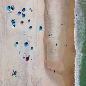 May include: An aerial view of a sandy beach with blue and white umbrellas, people swimming in the turquoise ocean, and white waves crashing on the shore.