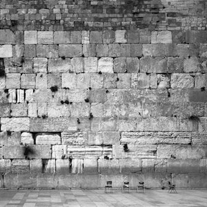 May include: A black and white photo of a stone wall with several chairs in front of it. The wall is made of large, rectangular stones, and the chairs are simple and wooden.