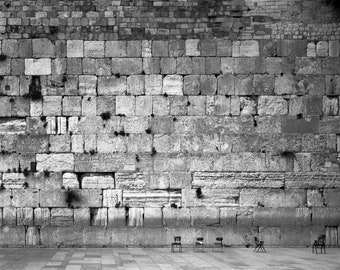 May include: A black and white photo of a stone wall with several chairs in front of it. The wall is made of large, rectangular stones, and the chairs are simple and wooden.