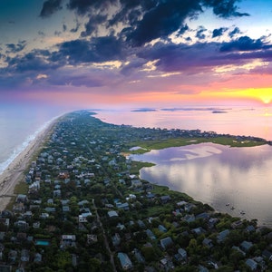 May include: Aerial view of a coastal landscape at sunset. The image shows a long, narrow strip of land with a beach on one side and a bay on the other. Houses and trees line the land. The sky is filled with colorful clouds, and the sun is setting over the water.