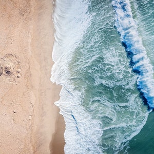 May include: An aerial view of a sandy beach with a lifeguard stand and a single person walking on the sand. The ocean waves are crashing on the shore, creating a white foamy line.
