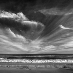May include: Black and white photograph of a beach scene. The sky is filled with dramatic, swirling clouds. The ocean waves crash onto the shore, and a wooden fence lines the beach.